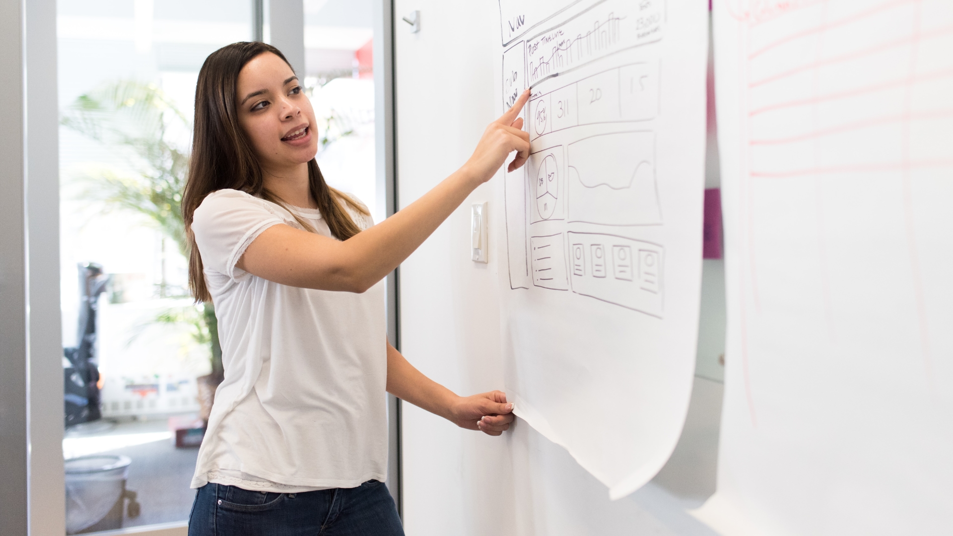 A woman at a whiteboard pointing to items in a wireframe sketch.
