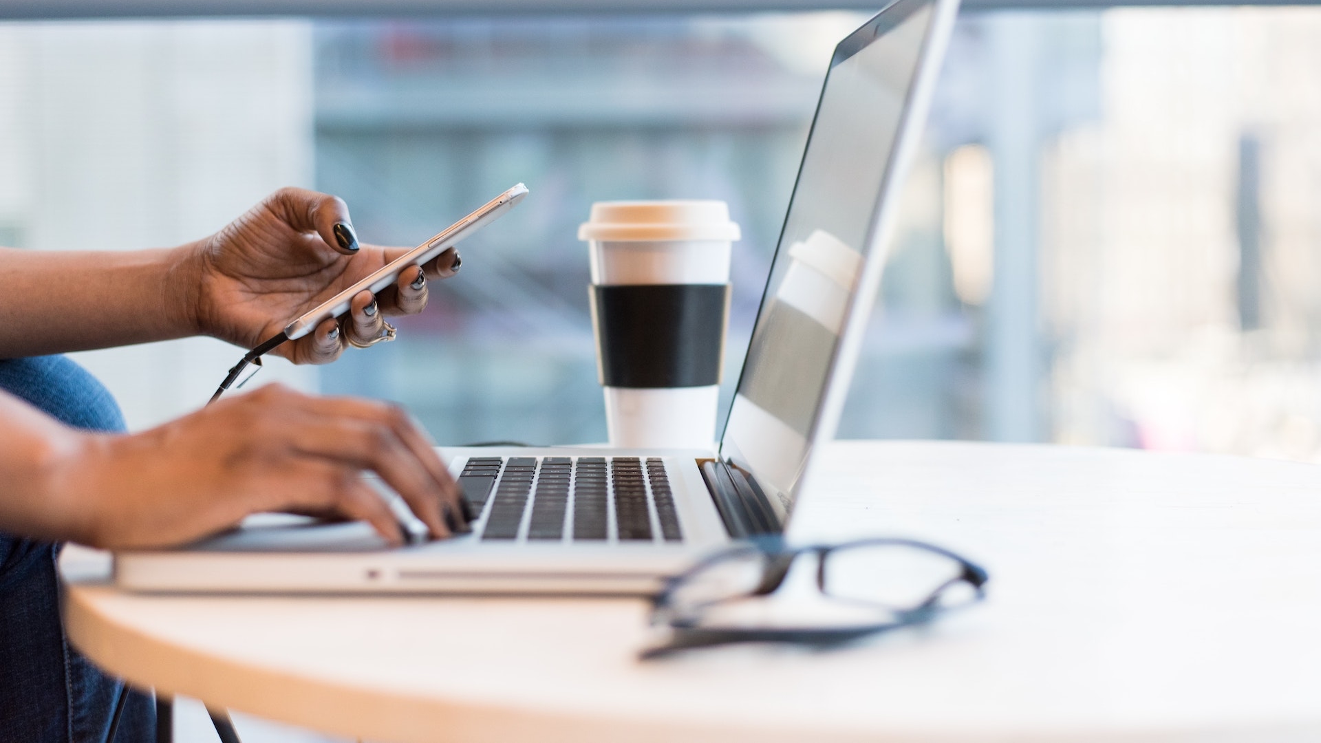 An African American woman’s hands holding an iPhone in the left hand and typing on a laptop keyboard with her right hand.