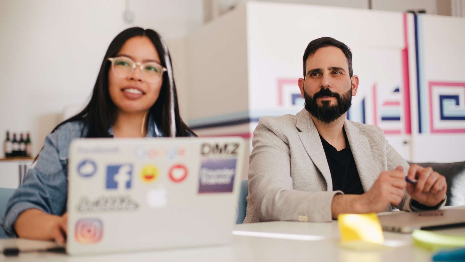 Asian woman and caucasian man sitting a meeting table. Their heads are turned towards the presenter.
