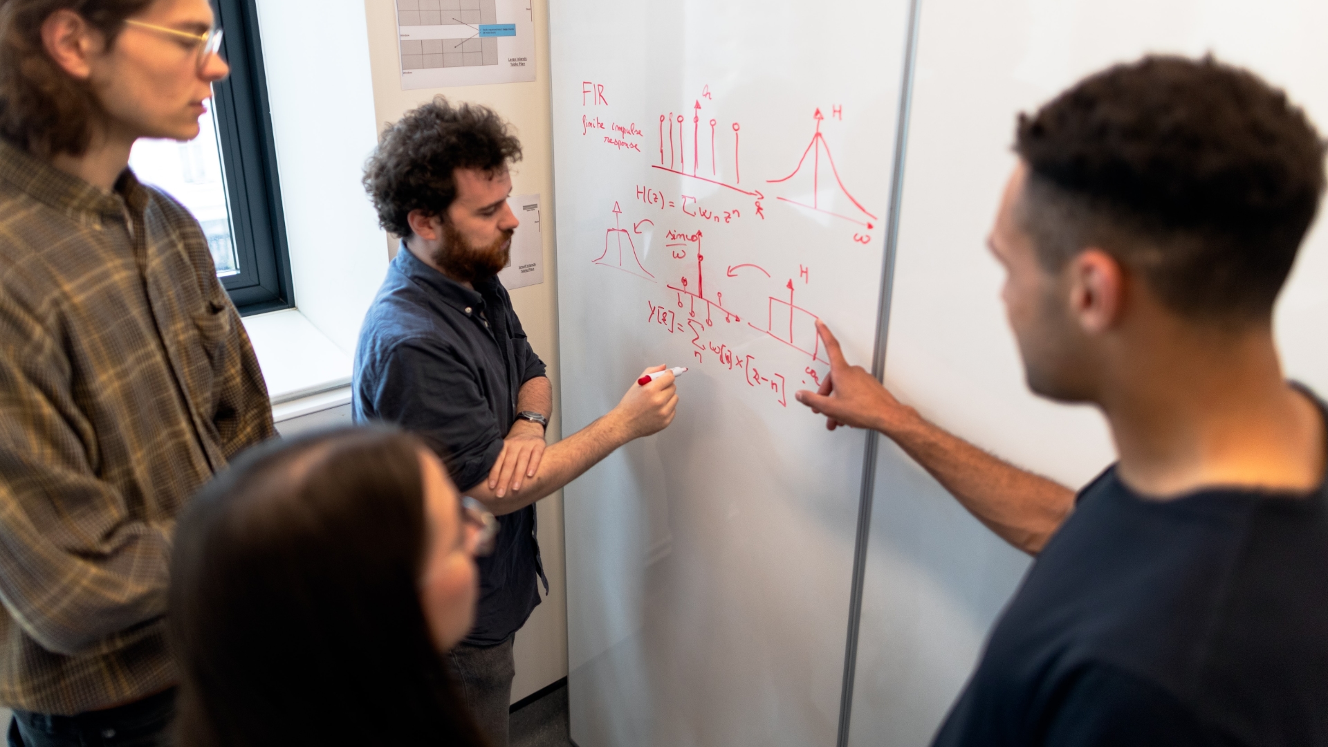 Four people, one of whom is a woman, pointing at math equations on a whiteboard.